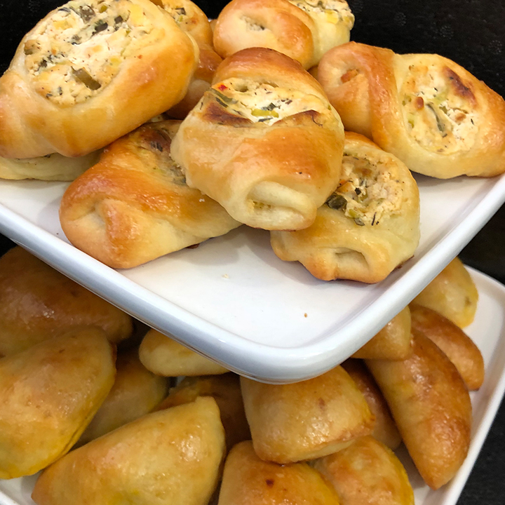 Frisch gebackene mit Schafskäse und Kräutern gefüllte Teigtaschen und Gebäckstücke auf zweistufigen weißen Servierplatten.